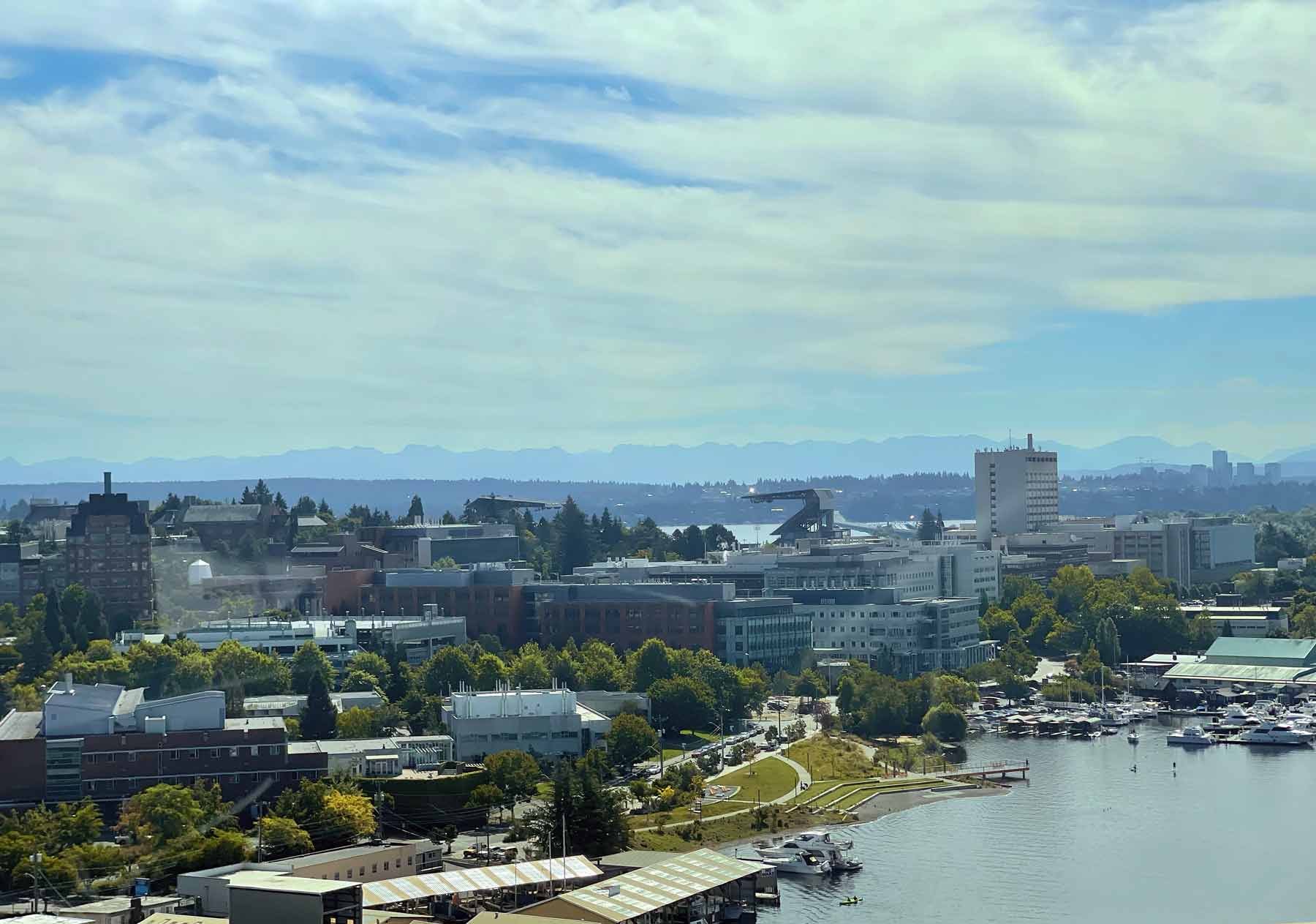 An elevated view of the University of Washington and the waterfront area, featuring a mix of modern buildings, green spaces, and a small marina with boats docked along the shore. The cityscape is set against a backdrop of distant mountains and a partly cloudy sky, adding depth to the expansive view. The setting feels lively yet calm, showcasing both the city's infrastructure and natural beauty.