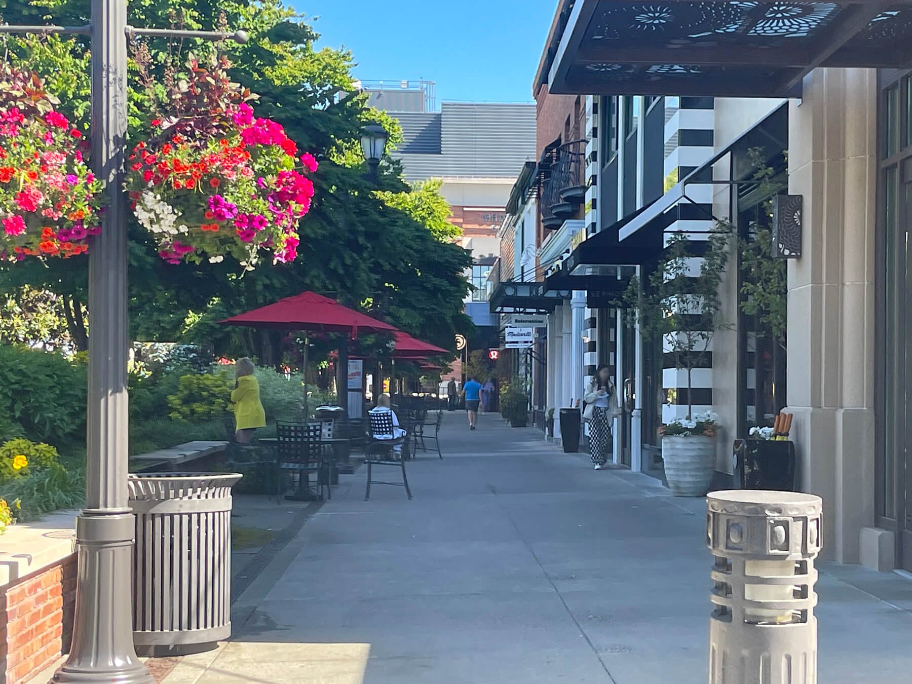 Pedestrian-friendly shopping street at U-Village in Seattle lined with boutique storefronts and outdoor seating under red umbrellas. Vibrant hanging flower baskets add a splash of color, while a few people stroll and enjoy the atmosphere. The scene is shaded by trees, creating a cozy and inviting urban vibe.