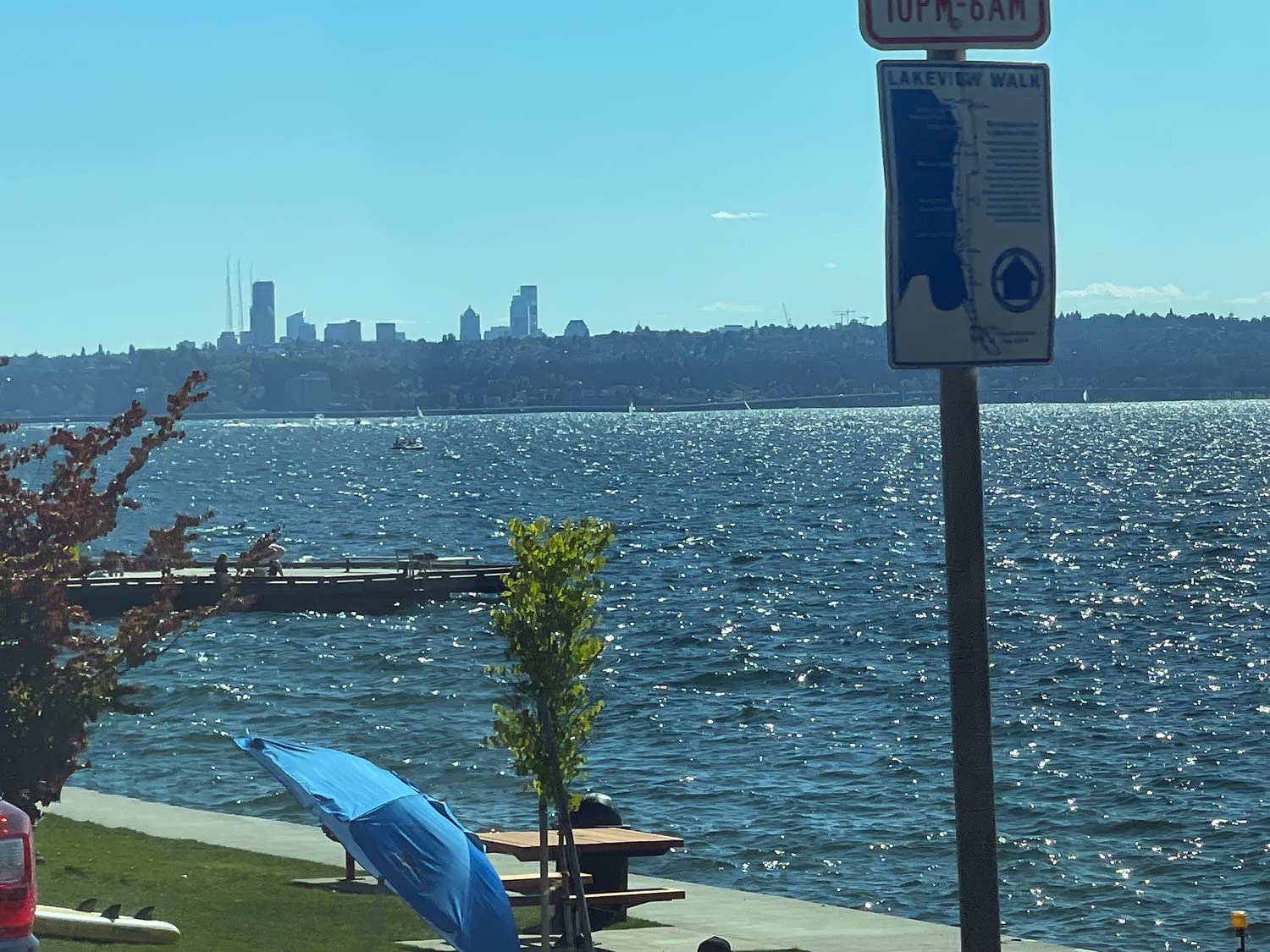 Kirkland Beach with scenic waterfront view on a sunny day with a clear blue sky. The Lakeview Walk sign is prominently displayed in the foreground, while the calm waters of the lake shimmer in the sunlight. A blue umbrella shades a picnic area along the grassy shore, and in the distance, small boats are seen gliding across the water. The far shoreline is dotted with trees and a city skyline, including prominent high-rise buildings of Seattle WA.

