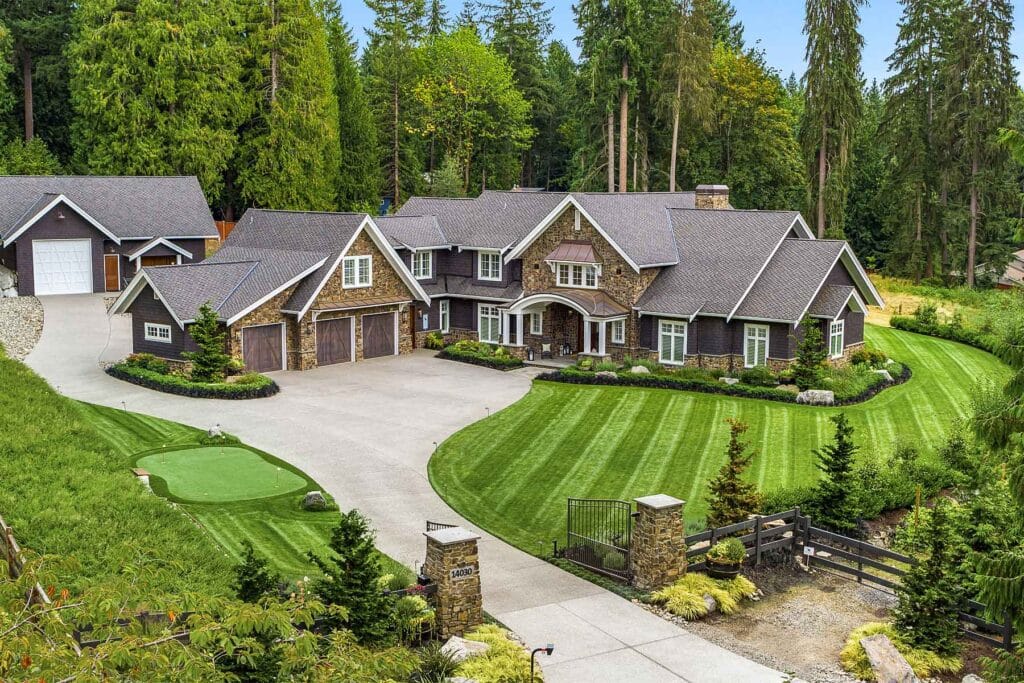 Aerial view of a luxurious, large country estate featuring a stone and dark wood exterior with white trim. The home has a grand entrance with a covered porch, multiple gables, and a spacious driveway leading to three garage doors. The meticulously manicured lawn includes a private putting green, and lush greenery surrounds the property, including tall evergreen trees. A detached garage or guesthouse is visible in the background, with a separate driveway curving up to it. A gated entrance with stone pillars adds an element of security and exclusivity to this expansive property nestled in a serene, wooded setting.